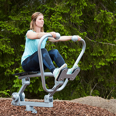 A woman exercises outdoors surrounded by nature. She is using an outdoor rowing machine.